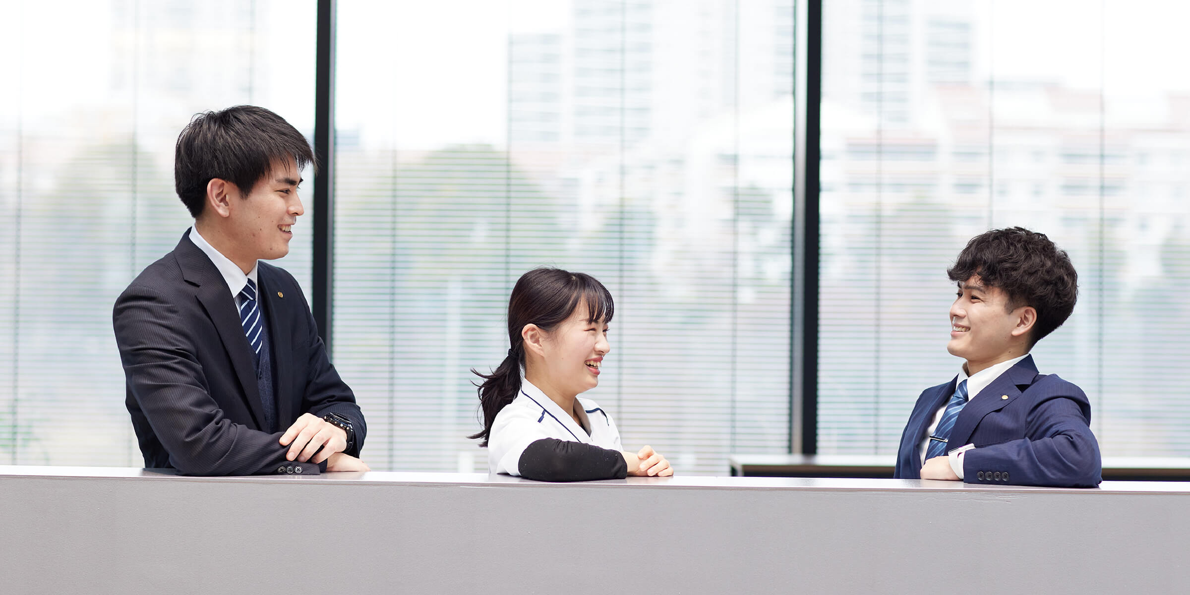 画像:3人、オフィスの棚の上に肘を乗せて、お話しているところ。すぐ後ろは窓際で、外には、木々とオフィスが見えている。