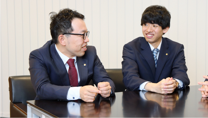 写真:スーツ姿の2人がテーブルに肘をつき、喋っている様子